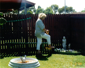 A woman in a blue shirt and white trousers, digging in a garden. Front left is a white garden feature with a chicken statue.