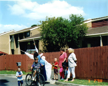 A group of women and children talking by the side of the road. One boy on a bicycle.