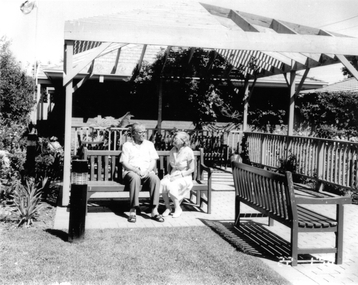 A man and a woman sitting on a park bench under a pergola.