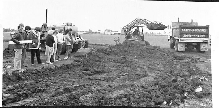 A group of people standing near a churned-up paddock watching a front-end loader loading a truck.
