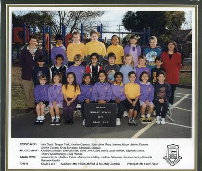 A group of children standing in three rows with teachers either side smiling at the camera with playground visible in background. 