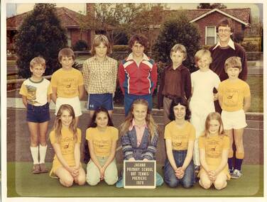 Two rows of children are positioned in the school playground. A teacher stands behind them in the top right-hand corner of the frame. Residential houses are visible in the background.