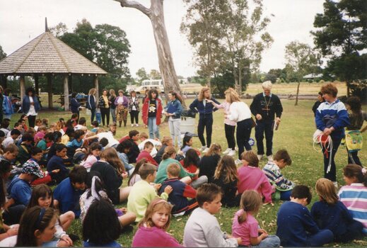 Children in rows, sitting on the Grass, in front of adults, with a pergola and trees behind them