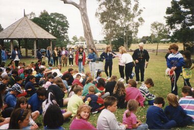 Children in rows, sitting on the Grass, in front of adults, with a pergola and trees behind them