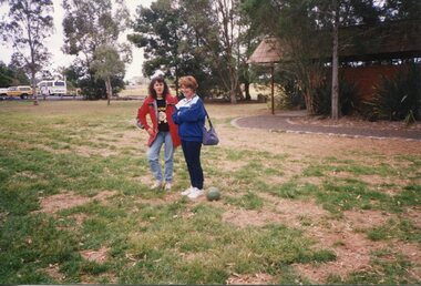 Two adults bin the park, with a brick building on the left in the background, and a carpark on the left
