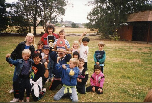 A group of young children posing for the camera in a park. A Brick building on the right in the background with trees