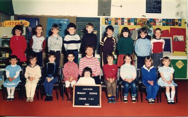 Formal class photograph, children in two rows, front row are seated on chairs. Centre of the front row seated is an adult, in front of whom is a child on the floor holding a board