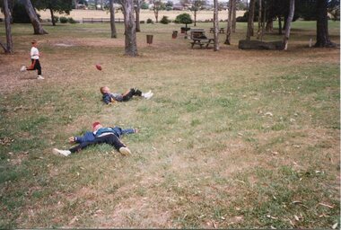 Children laying on the grass, laughing with a football in the air. One child is running in the background laughing