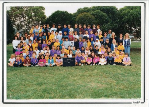 Photograph of children, in five rows, adults on each side of the third and fourth rows, with trees in the background