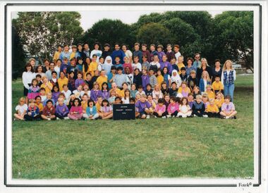 Photograph of children, in five rows, adults on each side of the third and fourth rows, with trees in the background