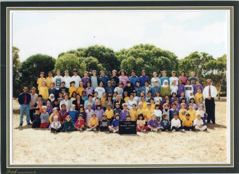 A Formal, posed, photograph of children in six rows and adults standing each end of the third row. There are trees in the background.