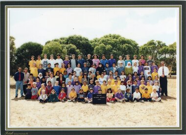 A Formal, posed, photograph of children in six rows and adults standing each end of the third row. There are trees in the background.