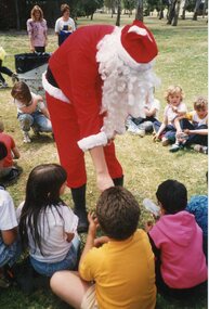 Father Christmas bending over to a child, seated on the grass. The child is part of a circle of children on the grass