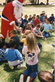 Father Christmas handing a wrapped item to a child. Children are sitting in a group on the grass.