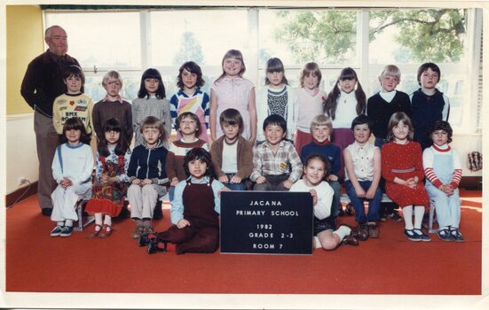 Formal photograph of three rows of children in front of windows. The front row of two children is sitting on the floor holding the title board. An adult is standing on the left behind the third row. 