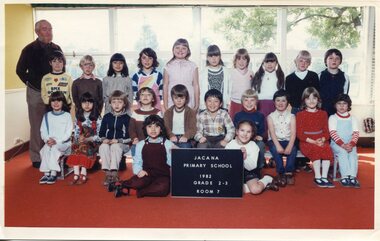 Formal photograph of three rows of children in front of windows. The front row of two children is sitting on the floor holding the title board. An adult is standing on the left behind the third row. 