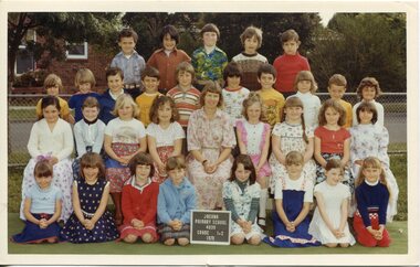 Formal photograph of children in four rows, with trees and houses in the background. The front row is kneeling on the ground. An adult is sitting centre of the second row. 