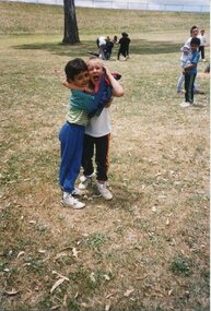 Two children centre hugging, with other children playing in the background