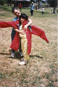 Two Children hugging, the one in front is wearing a red oversized coat. In the background are children playing.