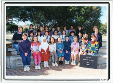 Formal Photograph of children sitting in three rows, on wooden benches, in front of trees. There is an adult sitting on each end of the second row
