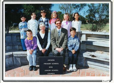 Photograph of children in two rows, the front row is sitting. In the middle of the front row are two seated adults. 