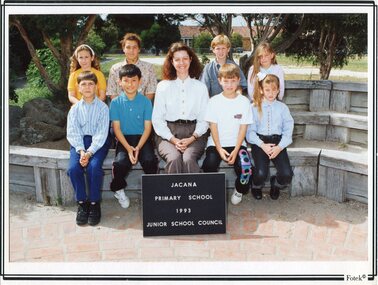 Formal Photograph of eight children, in two rows, sat on wooden seats. An adult is sat centre of the front row. 