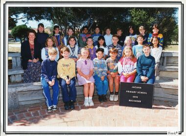 Formal Photograph of Children, sitting in three Rows on wooden seats. An adult is sitting on the left of the second row.