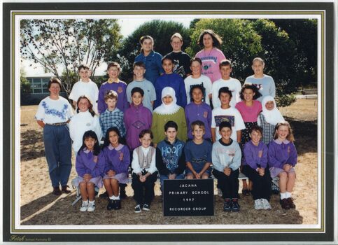 Formal Group photograph with children in four rows, with tress and buildings in the background. An adult is standing to the left of the second row.  