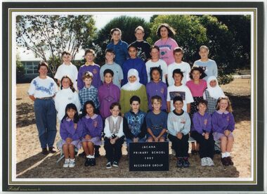 Formal Group photograph with children in four rows, with tress and buildings in the background. An adult is standing to the left of the second row.  