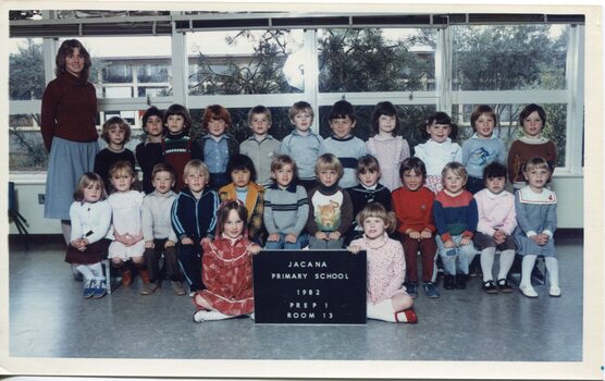Formal photograph of children in three rows, in a classroom. An Adult is standing on the left in the back row. 