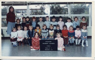 Formal photograph of children in three rows, in a classroom. An Adult is standing on the left in the back row. 