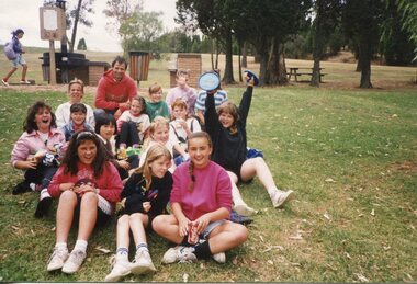Group of children sitting on the grass in front of a sign and some trees.