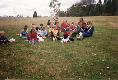 A group of Children sitting on the grass, with two adults on the right