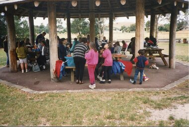 Children and adults beneath a wooden pergola at wooden tables and benches