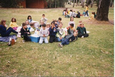 Group of children sitting on the grass eating snack with a brick building and trees in the background