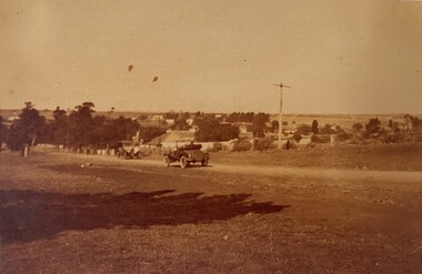Two cars driving on a dirt road, with houses in the background