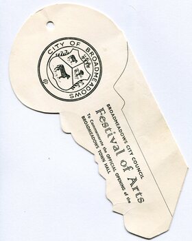Front of the die-cut programme showing the City of Broadmeadows coat of arms printed in black on cream paper. Adjacent text reads “Broadmeadows City Council Festival of Arts” and references the official opening of the Broadmeadows Town Hall. The object has a curved, fan-like outline with a central fold and a suspension hole near the top.
