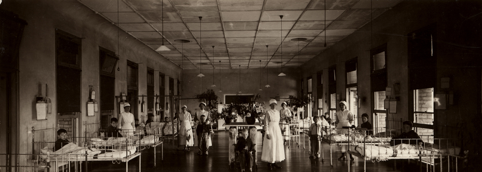 Ormond Ward, circa 1920. Long shot of interior of hospital room. Beds are positioned along walls, staff are present throughout the room while there are patients standing, sitting and in beds.