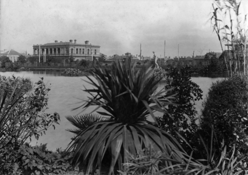 Sir Redmond Barry's house from Carlton Gardens, 1876. A long shot of Carlton Gardens, shrubs in the foreground a lake in the middle and buildings  in the background.
