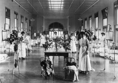 Snowball Ward, Children's Hospital, Carlton, 1908. A long shot of a ward area with approximately ten hospital cots with children either laying down or sitting right and left of camera. Seven staff members in uniform are standing alongside the cots. Two children sitting in chairs in front of a table with vases of flowers are in foreground.