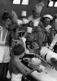 The birthday party, The Children's Hospital Orthopaedic Section, circa 1937. A medium shot of a group of young patients and a Nurse standing around a metal framed bed. The children are wearing paper hats, have drinks and are celebrating the birthday of a young child, wearing leg frames and an arm brace, lying on the bed with a birthday cake beside them.
