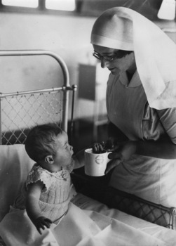 Nurse and infant patient at the Children's Hospital Orthopaedic Section, Mt Eliza, circa 1947. A mid shot of an infant sitting on a hospital bed, a staff member in uniform standing beside the bed is holding a large mug.