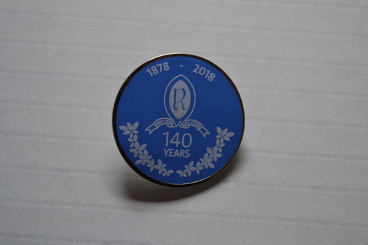 Circular metal lapel badge with mid-blue and white decoration of a school crest.