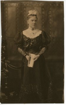 A older woman standing in a photographic studio.  She is wearing a long dark dress and holds a book in front of her.