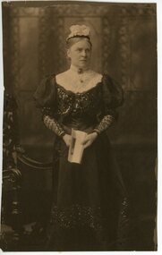 A older woman standing in a photographic studio.  She is wearing a long dark dress and holds a book in front of her.