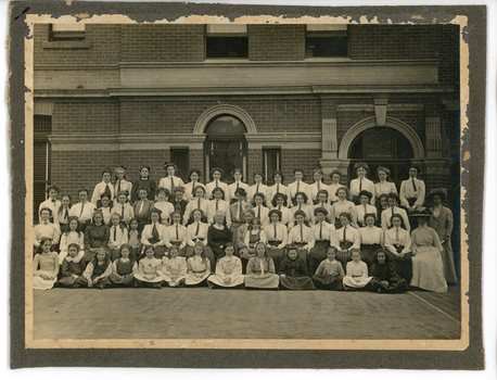 Black and white group photo of adult women and a large group of children set in front of a brick building.