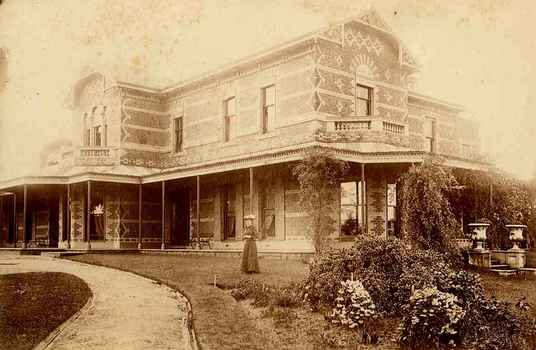 Sepia photo of woman standing in a garden in front of two story brick house.