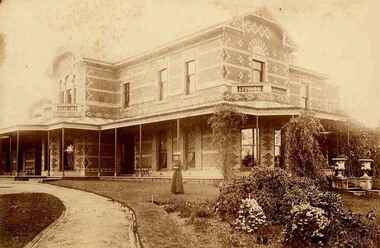 Sepia photo of woman standing in a garden in front of two story brick house.