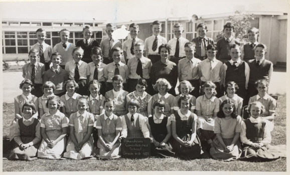 Four rows of children standing outside in front of a school. Their teacher is in the middle of the students, and one student on the bottom row is holding up a sign stating 'Pakenham Consolidated School Grade VI b 1957