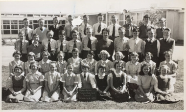 Four rows of children standing outside in front of a school. Their teacher is in the middle of the students, and one student on the bottom row is holding up a sign stating 'Pakenham Consolidated School Grade VI b 1957